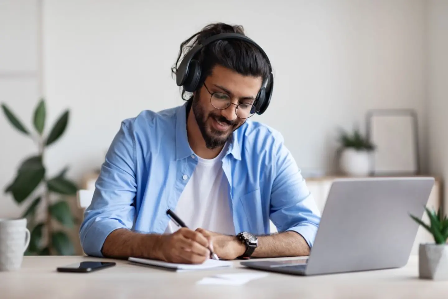 Man wearing headphones studying online at laptop and taking notes.